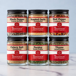 Six glass spice jars arranged in two rows of three on a light marble surface against a pale blue-gray background. The jars are labeled: Black Pepper Coarse Ground, Roasted Garlic Granulated, Red Pepper Crushed Flakes, Celery Salt Granulated, Parsley Dried Flakes, and Thyme Dried Flakes, all by Tradition Spice Company.