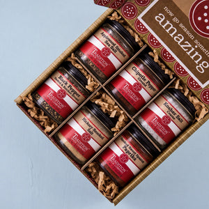 An open gift box of eight Trader Joe's seasoning jars arranged in a grid on a light blue background. The box is lined with brown paper shred and has a brown header card that reads 'now go season something amazing'.