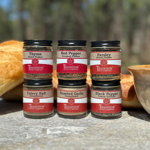 Five jars of spices from Tradition Spice Company on a wooden surface with bread and a bowl in the background.