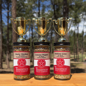 The American Kitchen Mega Pack from Tradition Spice Company features three citrus spice blends—Zesty Chicken, Lemony Pepper, and Sublime Lime—displayed outdoors on wood with gold trophies, perfect for any weeknight meal. Pine trees blur behind.
