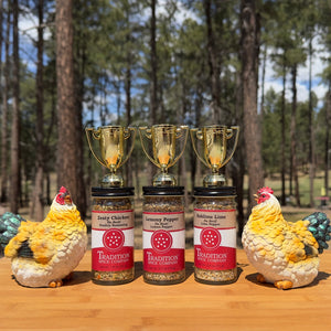 The American Kitchen Mega Pack, ideal for chicken seasoning or citrus spice blends, features three spice jars displayed outdoors on a wooden surface with gold trophies and ceramic chickens amid tall pine trees.